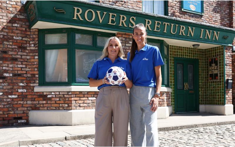 Jill Scott stands with a woman outside the Rover's Return on the Coronation Street set. the other woman is holing a football. Both women wear National Lottery blue polo shirts