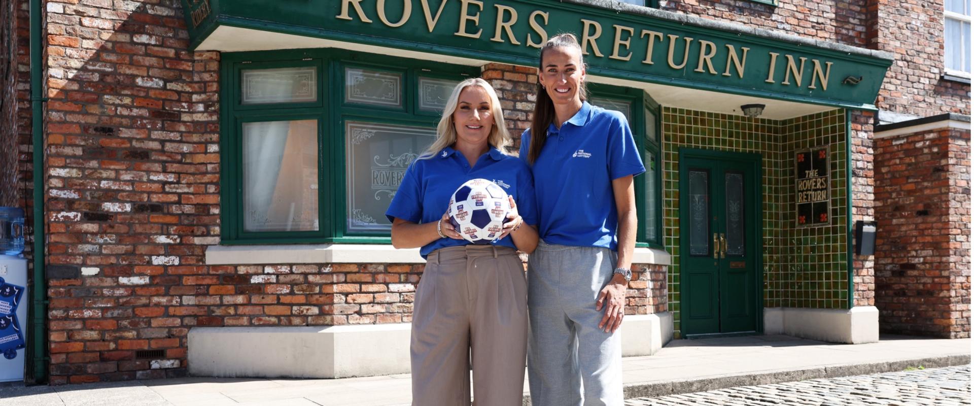 Jill Scott stands with a woman outside the Rover's Return on the Coronation Street set. the other woman is holing a football. Both women wear National Lottery blue polo shirts