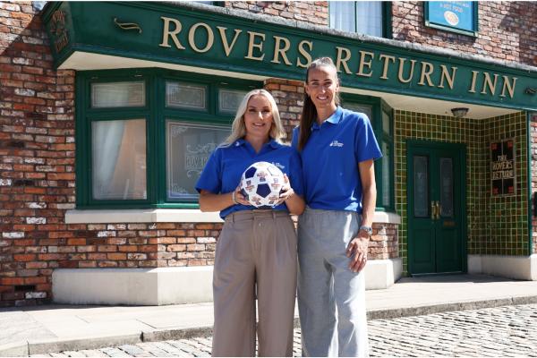 Jill Scott stands with a woman outside the Rover's Return on the Coronation Street set. the other woman is holing a football. Both women wear National Lottery blue polo shirts