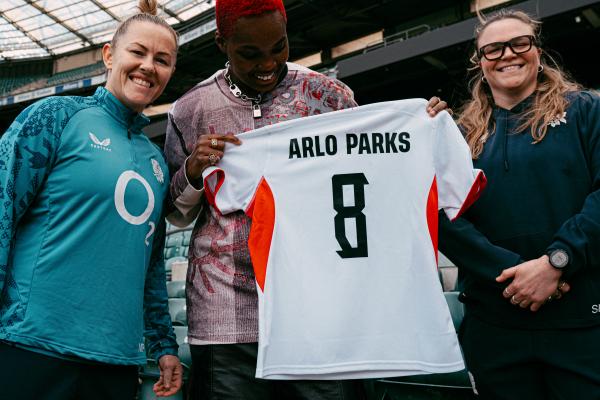 Arlo parks with Marlie Packer and Sarah Bern at Twickenham holding a red roses shirt with ARLO PARKS 8 on the back. they are all smiling and looking at the shirt