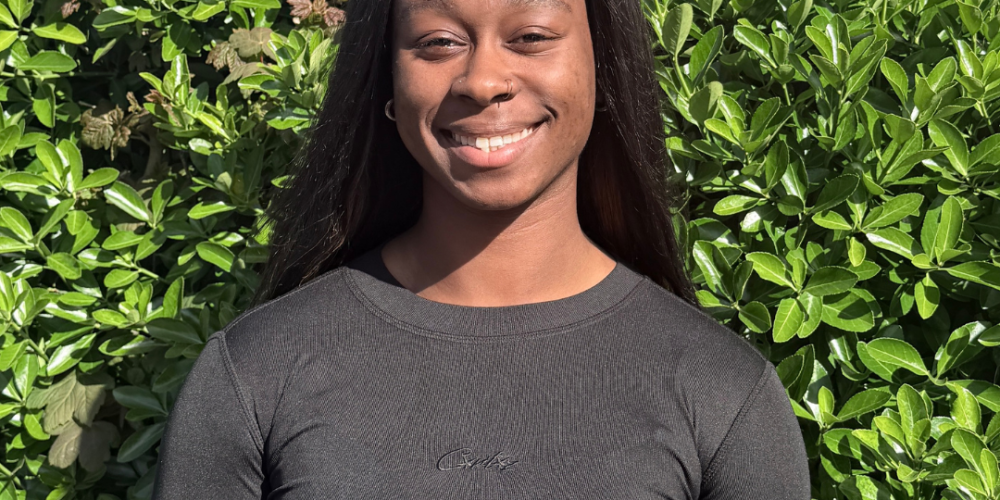 Chante stands in front of a green bush. there is blue sky in teh top left corner of the image and a brick building to the right. She is wearing all black and is smiling. She has long black hair that sits behind her shoulders
