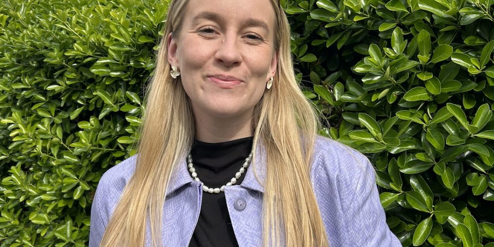 Ellen stands in front of a green bush and cloudy sky. She has blonde hair with sunglasses on top of her head. She is smiling. She wears a lilac jacket and a black top with a pearl necklace on.