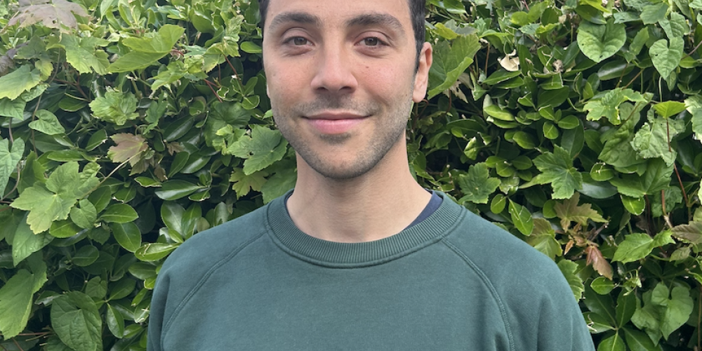 Nathan stands in front of a green hedge. he is smiling. He is wearing a green jumper with the words parra on it. He has dark hair 