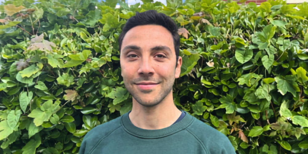 Nathan stands in front of a green hedge. he is smiling. He is wearing a green jumper with the words parra on it. He has dark hair 